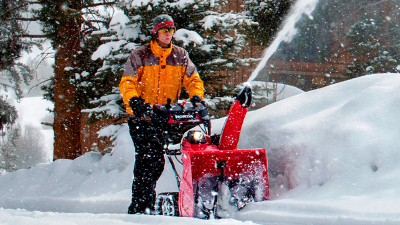 Wide view of a man clearing a bath with a snowblower just outside a chalet.